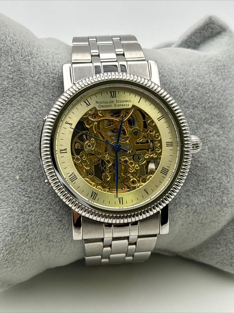 May include: Silver-toned wristwatch with a skeleton dial, showcasing the intricate inner workings. The watch features Roman numeral hour markers and the text "Nostalgie Istanbul Orient Express" on the face. The watch band is made of linked metal.