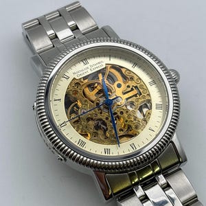 May include: A silver-toned wristwatch with a skeleton dial, showcasing the intricate inner workings. The watch features Roman numeral hour markers and blue hands. The text "Nostalgie Istanbul Orient Express" is visible on the watch face. The watch band is made of silver-colored metal.