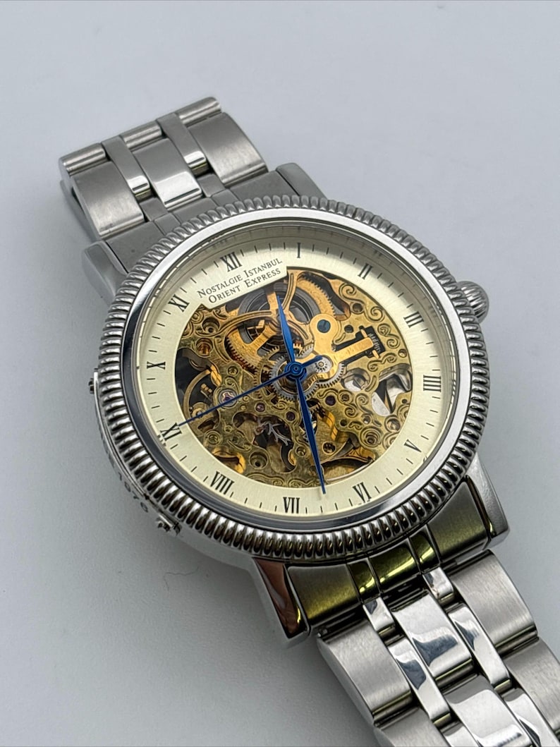 May include: A silver-toned wristwatch with a skeleton dial, showcasing the intricate inner workings. The watch features Roman numeral hour markers and blue hands. The text "Nostalgie Istanbul Orient Express" is visible on the watch face. The watch band is made of silver-colored metal.