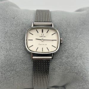 Pode incluir: Um relógio Omega Genève vintage com uma pulseira de malha prateada. O mostrador é branco com marcadores de horas e ponteiros pretos. O logotipo da Omega e "Genève" estão impressos no mostrador. O relógio tem uma caixa retangular prateada.