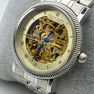 May include: Silver-toned wristwatch with a skeleton dial, showcasing the intricate inner workings. The watch features Roman numeral hour markers and blue hands. The band is made of linked metal. The watch face reads "Nostalgie Istanbul Orient Express".