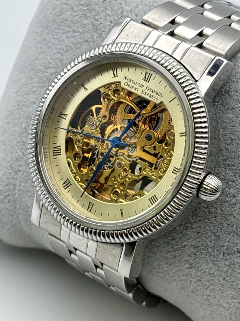 May include: Silver-toned wristwatch with a skeleton dial, showcasing the intricate inner workings. The watch features Roman numeral hour markers and blue hands. The band is made of linked metal. The watch face reads "Nostalgie Istanbul Orient Express".