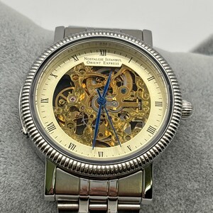 May include: A silver-toned wristwatch with a skeleton dial, showcasing the intricate inner workings. The watch features Roman numeral hour markers and the text "Nostalgie Istanbul Orient Express" on the face. The watchband is also silver-toned.