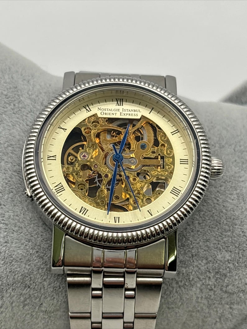 May include: A silver-toned wristwatch with a skeleton dial, showcasing the intricate inner workings. The watch features Roman numeral hour markers and the text "Nostalgie Istanbul Orient Express" on the face. The watchband is also silver-toned.