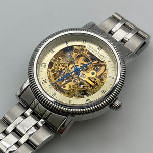 May include: A silver-toned wristwatch with a skeleton dial, showcasing the intricate inner workings. The watch features Roman numeral hour markers and blue hands. The band is made of linked metal. The watch face reads "Nostalgie Istanbul Orient Express."