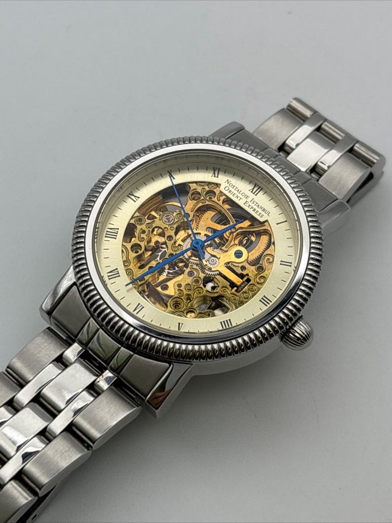 May include: A silver-toned wristwatch with a skeleton dial, showcasing the intricate inner workings. The watch features Roman numeral hour markers and blue hands. The band is made of linked metal. The watch face reads "Nostalgie Istanbul Orient Express."