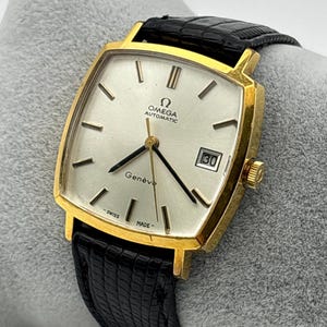 Pode incluir: Um relógio de pulso automático Omega Genève dourado, com mostrador quadrado e bracelete de couro preto. O mostrador prateado tem ponteiros e marcadores pretos, e uma janela de data às 3 horas. O relógio está marcado "Swiss Made".