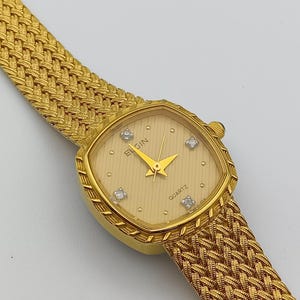 Puede incluir: Un reloj Elgin de cuarzo dorado con esfera cuadrada y correa trenzada. La esfera del reloj es de color beige claro con cuatro pequeños diamantes. Las manecillas del reloj son doradas y tienen forma de flecha. La correa es de diseño trenzado dorado a juego.