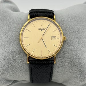 Könnte beinhalten: Eine goldfarbene Longines-Uhr mit einem schwarzen Lederarmband. Das Zifferblatt der Uhr ist goldfarben mit goldenen Zeigern und Markierungen sowie einem Datumsfenster bei 3 Uhr. Die Uhr wird auf einer grauen Oberfläche präsentiert.