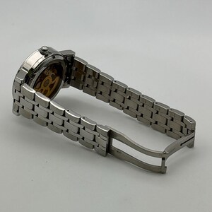 May include: A silver-toned wristwatch with a stainless steel band. The watch face is visible, showcasing the intricate inner workings. The band is open, displaying the clasp mechanism. The watch is set against a plain white background.