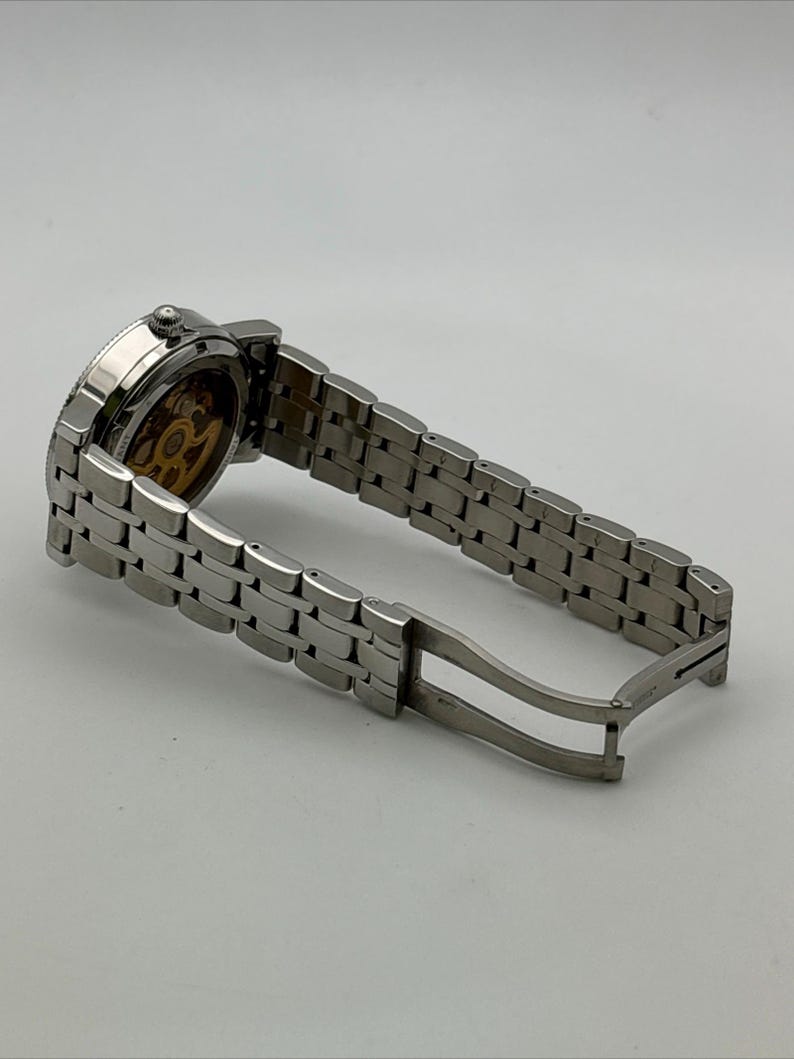 May include: A silver-toned wristwatch with a stainless steel band. The watch face is visible, showcasing the intricate inner workings. The band is open, displaying the clasp mechanism. The watch is set against a plain white background.