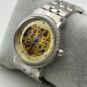 May include: A silver-toned wristwatch with a skeleton dial, showcasing the intricate inner workings. The watch features Roman numeral hour markers and the text "Nostalgie Istanbul Orient Express". The watch is on a silver metal band.