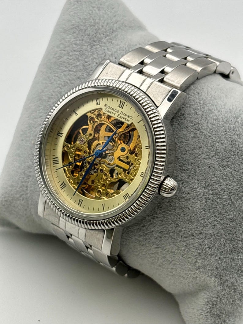 May include: A silver-toned wristwatch with a skeleton dial, showcasing the intricate inner workings. The watch features Roman numeral hour markers and the text "Nostalgie Istanbul Orient Express". The watch is on a silver metal band.