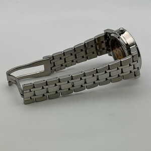 May include: A silver-toned wristwatch with a stainless steel band. The watch face is visible, showcasing intricate internal mechanisms. The band is open, displaying the clasp. The watch is a luxury item.