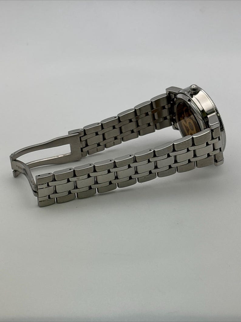 May include: A silver-toned wristwatch with a stainless steel band. The watch face is visible, showcasing intricate internal mechanisms. The band is open, displaying the clasp. The watch is a luxury item.
