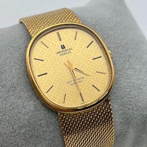 May include: A gold-toned Universal Geneve Gilt Shadow automatic wristwatch. The watch has a rectangular face with a patterned design and black markers. The watchband is a mesh style in a matching gold tone.