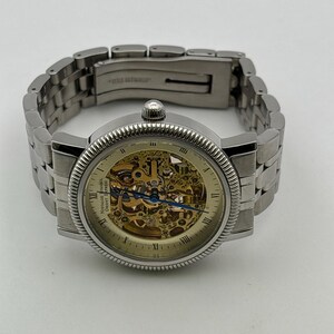 May include: A silver-toned wristwatch with a skeleton dial, showcasing the intricate inner workings. The watch features Roman numeral hour markers and a textured bezel. The stainless steel band is connected with a clasp. The watch is a classic design.