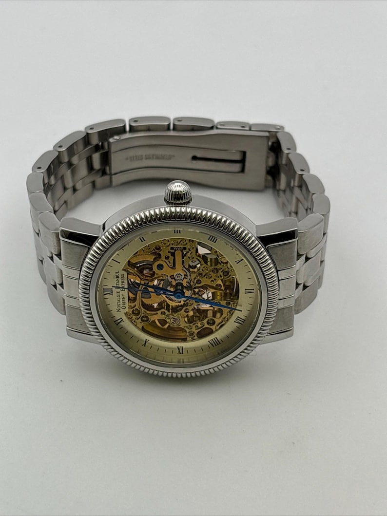 May include: A silver-toned wristwatch with a skeleton dial, showcasing the intricate inner workings. The watch features Roman numeral hour markers and a textured bezel. The stainless steel band is connected with a clasp. The watch is a classic design.