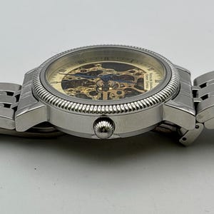 May include: A silver-toned wristwatch with a skeleton dial, showcasing the intricate inner workings. The watch features a textured bezel and a linked metal band. The dial has gold and blue accents, and the watch is set against a white background.