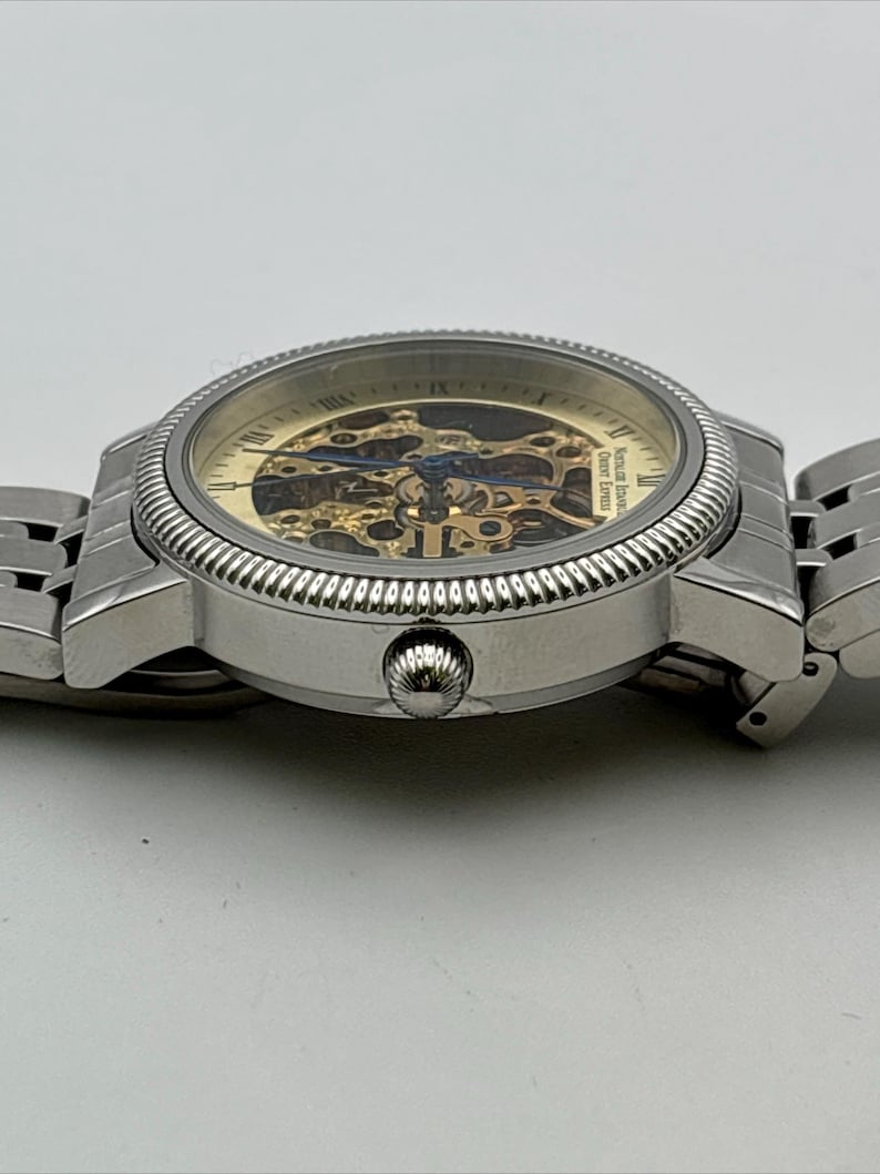 May include: A silver-toned wristwatch with a skeleton dial, showcasing the intricate inner workings. The watch features a textured bezel and a linked metal band. The dial has gold and blue accents, and the watch is set against a white background.