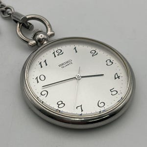 Op de afbeelding: Een zilveren Seiko Quartz zakhorloge met een witte wijzerplaat en zwarte cijfers. Het horloge heeft een ketting en een ronde, gepolijste kast. De wijzerplaat toont het Seiko Quartz logo.