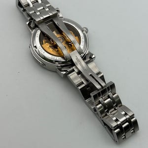 May include: A silver stainless steel wristwatch with a transparent back, revealing the intricate gold-colored internal mechanisms. The watch has a linked band and is displayed against a light background.