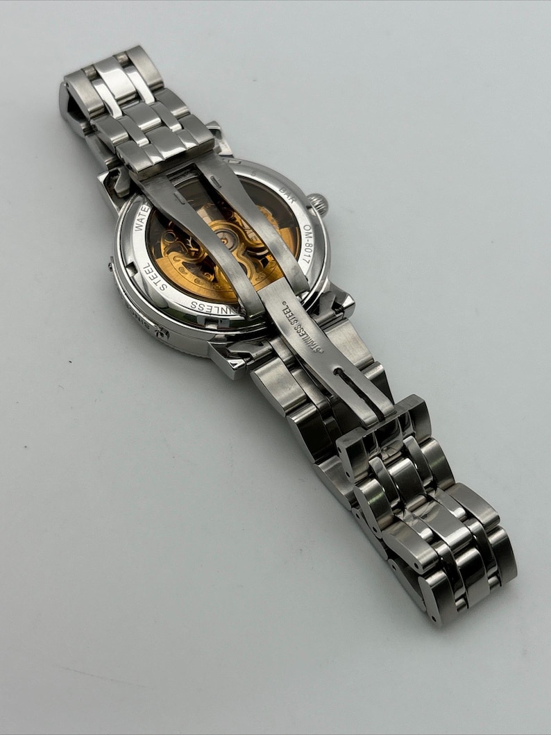 May include: A silver stainless steel wristwatch with a transparent back, revealing the intricate gold-colored internal mechanisms. The watch has a linked band and is displayed against a light background.