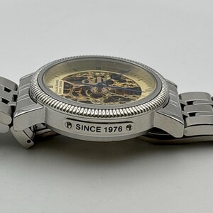 May include: A silver-toned wristwatch with a skeleton dial, showcasing the intricate inner workings. The watch features a textured bezel and a metal band. The text "SINCE 1976" is visible on the side of the watch.