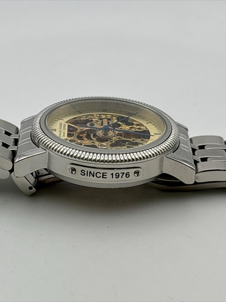 May include: A silver-toned wristwatch with a skeleton dial, showcasing the intricate inner workings. The watch features a textured bezel and a metal band. The text "SINCE 1976" is visible on the side of the watch.