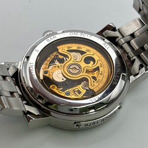 May include: A silver stainless steel wristwatch with a transparent back, showcasing the intricate gold-colored mechanical movement. The watch features the text "OM-8017" and "Since 1976". The watch band is also silver stainless steel.