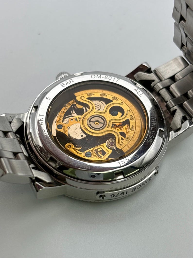 May include: A silver stainless steel wristwatch with a transparent back, showcasing the intricate gold-colored mechanical movement. The watch features the text "OM-8017" and "Since 1976". The watch band is also silver stainless steel.
