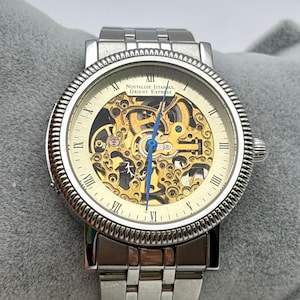 May include: A silver-toned wristwatch with a skeleton dial, showcasing the intricate inner workings. The watch features Roman numeral hour markers and a blue second hand. The watch band is made of silver links, and the watch face reads "Nostalgie Istanbul Orient Express."