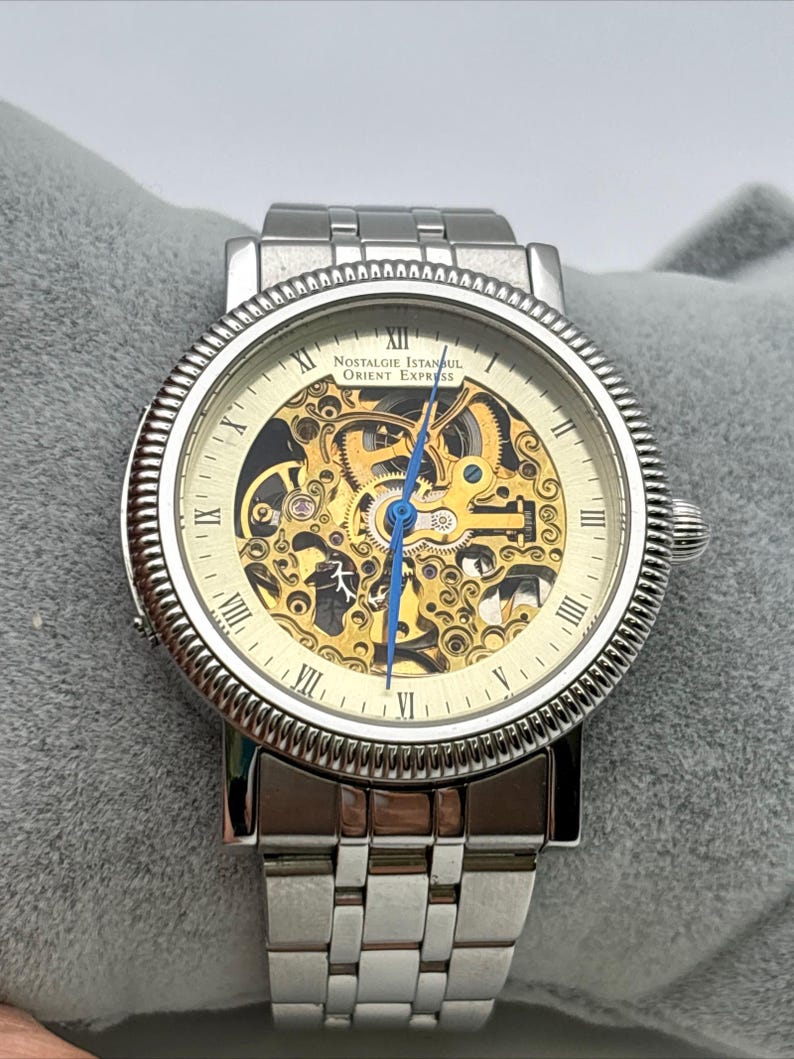 May include: A silver-toned wristwatch with a skeleton dial, showcasing the intricate inner workings. The watch features Roman numeral hour markers and a blue second hand. The watch band is made of silver links, and the watch face reads "Nostalgie Istanbul Orient Express."