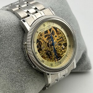 May include: Silver-toned wristwatch with a skeleton dial, showcasing the intricate inner workings. The watch features Roman numeral hour markers and a silver link band. The dial reads "Nostalgie Istanbul Orient Express".
