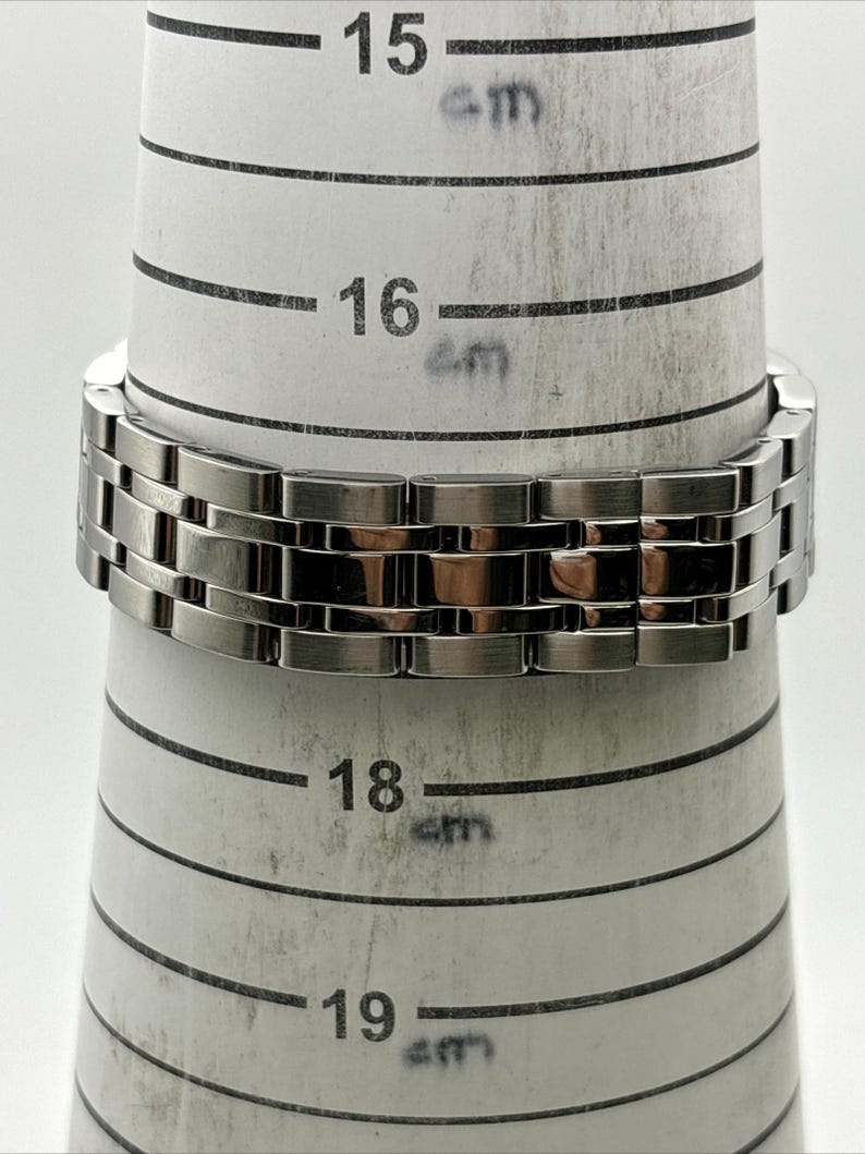 May include: A silver-toned metal bracelet is wrapped around a ring sizer. The bracelet is made of rectangular links. The ring sizer has measurements in centimeters, with the bracelet resting at the 16 cm mark.