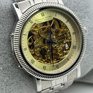 May include: Silver-toned wristwatch with a skeleton dial, showcasing the intricate inner workings. The watch features Roman numeral hour markers and the text "Nostalgie Istanbul Orient Express". The watch has a silver band.