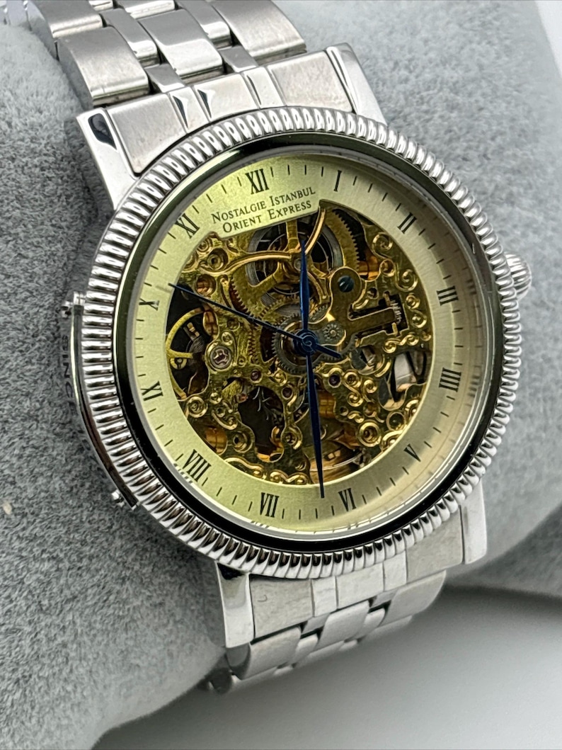 May include: Silver-toned wristwatch with a skeleton dial, showcasing the intricate inner workings. The watch features Roman numeral hour markers and the text "Nostalgie Istanbul Orient Express". The watch has a silver band.