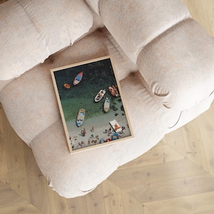 May include: A framed print featuring a beach scene with several boats floating on turquoise water near a sandy shore. People are gathered on the beach. The print is resting on a beige upholstered chair.