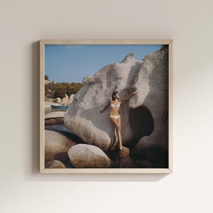 Könnte beinhalten: Gerahmter Druck mit einer Frau in einem weißen Bikini, die in der Nähe einer großen, hellfarbigen Felsformation posiert. Die Szene spielt an einem Strand mit klarem blauem Himmel und einem kleinen Boot im Hintergrund. Das Kunstwerk ist in einem hellfarbigen Holzrahmen eingefasst.