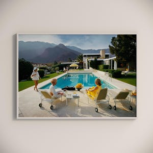 May include: A framed print depicts a retro scene by a pool. Two women in stylish attire relax on lounge chairs, while another walks nearby. The pool is surrounded by a patio, lush greenery, and mountains in the background.