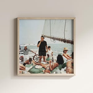 May include: A framed print depicts a vintage scene of people enjoying a yacht outing. The image shows a group of people on a boat, with a man smoking, others lounging, and a child. The scene is set against a backdrop of the sea and sky.
