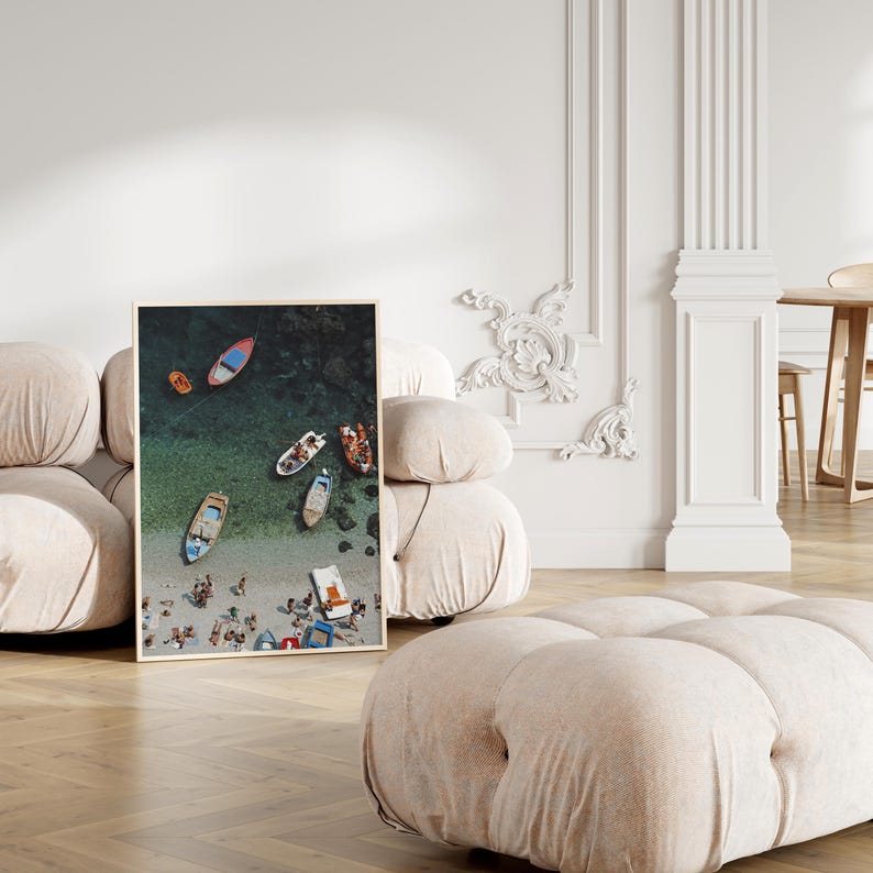 May include: A framed print displays an aerial view of a beach scene. Several colorful boats are docked near the shore, with people and beach umbrellas visible on the sandy beach. The water transitions from turquoise to deep blue.