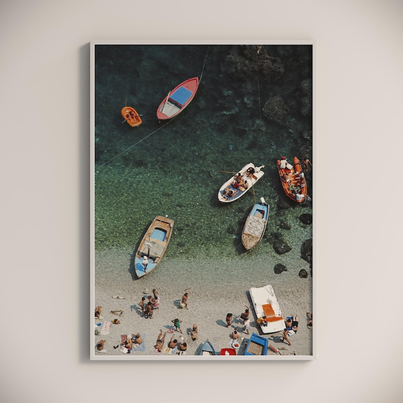 May include: Aerial view of a beach scene with several small boats floating in turquoise water. People are sunbathing on the sandy shore. The boats are various colors, including red, orange, and blue. The image is framed in a white border.
