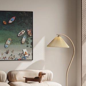 May include: A framed aerial photograph of a beach scene with several colorful boats and people. The photograph is displayed on a wall above a beige sofa with a dachshund figurine. A gold floor lamp with a pleated shade is to the right.