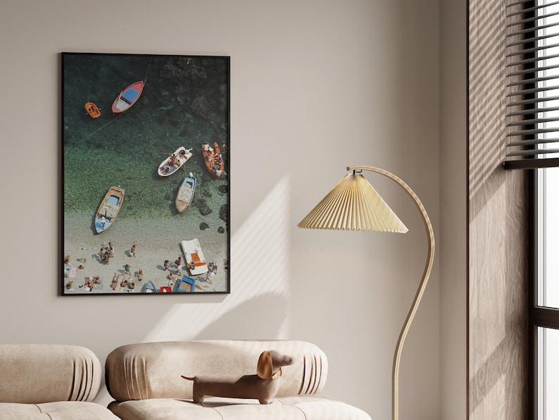 May include: A framed aerial photograph of a beach scene with several colorful boats and people. The photograph is displayed on a wall above a beige sofa with a dachshund figurine. A gold floor lamp with a pleated shade is to the right.