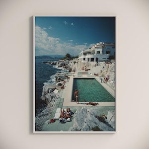 May include: A framed print depicts a coastal scene with a turquoise pool, white buildings, and people enjoying the sun. The image captures a sunny day with a clear blue sky and the Mediterranean Sea. The print evokes a sense of leisure and travel.