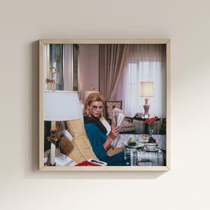 May include: A framed print of a woman in a blue dress reading a newspaper, with a decorative eye mask. The scene is set in a room with a lamp, curtains, and a table with food and drinks. The print has a light wood frame.