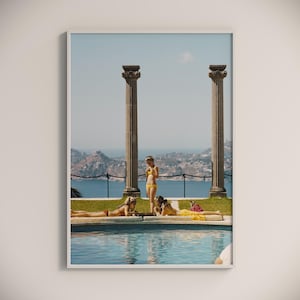 May include: Four women in swimsuits by a pool with a view of a distant town and ocean. Two women are laying by the pool, while two women stand by the edge of the pool. The pool is surrounded by a stone wall with columns.