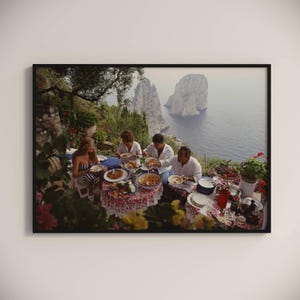 Puede incluir: Un grupo de personas están sentadas alrededor de una mesa al aire libre, disfrutando de una comida con vistas al océano y una isla rocosa en la distancia. La mesa está cubierta con un mantel colorido y hay platos de comida en la mesa. Las personas están sonriendo y riendo.