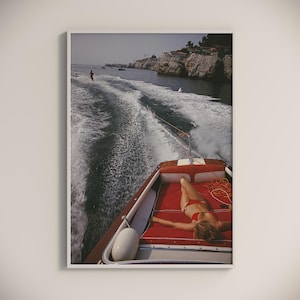 Op de afbeelding: Een ingelijste print van een vintage foto met een waterskiër die door een boot wordt getrokken. Een persoon in een rode bikini zonnebaadt op het rood gevoerde dek van de boot. De boot beweegt door het water en creëert een kielzog.